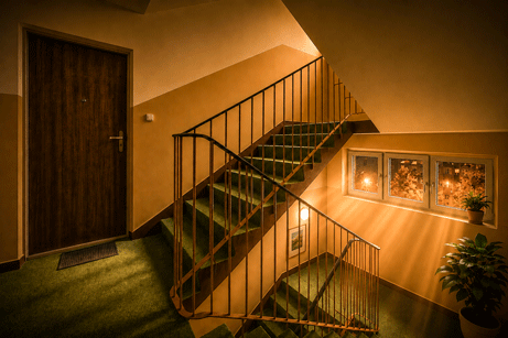 A  night scene of an apartment stairwell and stairs. Light pours from the tree windows from nearby streetlamps spill onto the green carpet on the stairwell. 